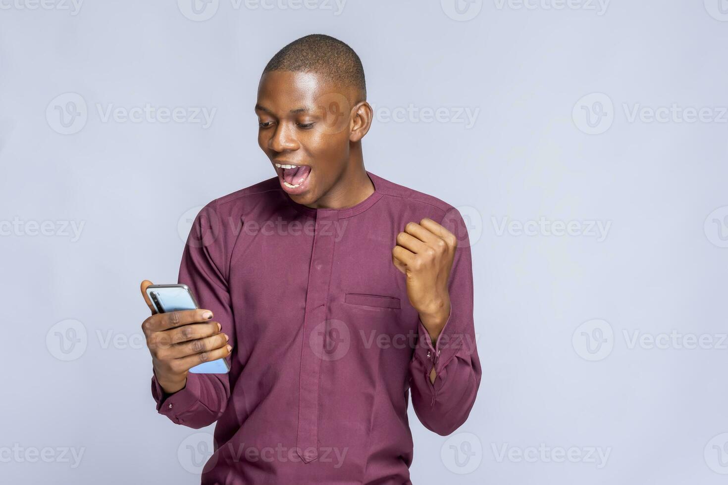 happy joyful man holds mobile phone and celebrates win makes fist bump laughs has surprised expression wears africa clothing stands sideways against white background empty space photo