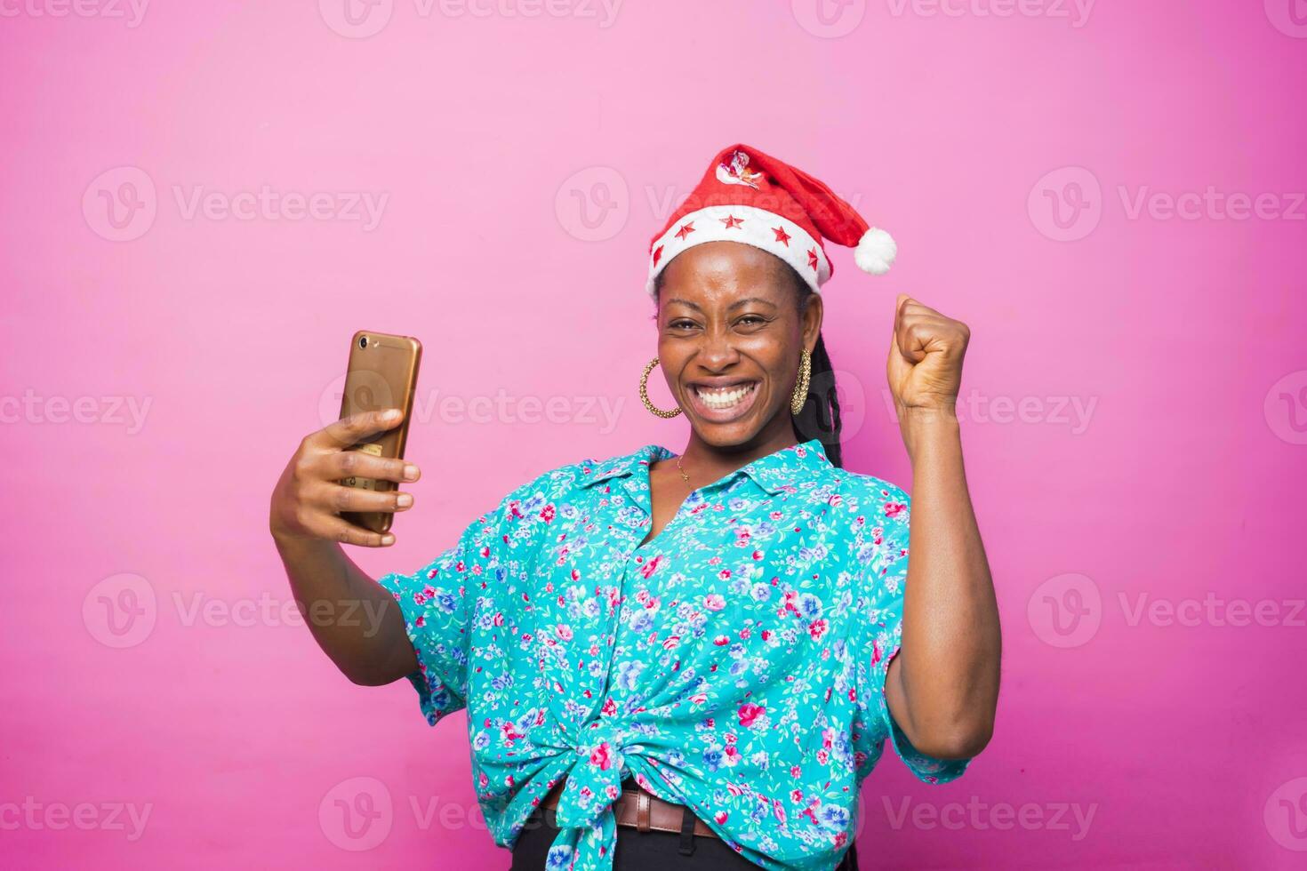 sobreexcitado negro africano hembra milenario celebrando con su inteligente teléfono después recepción bueno noticias. vistiendo Navidad sombrero en pie detrás un rosado estudio pared. Navidad concepto foto