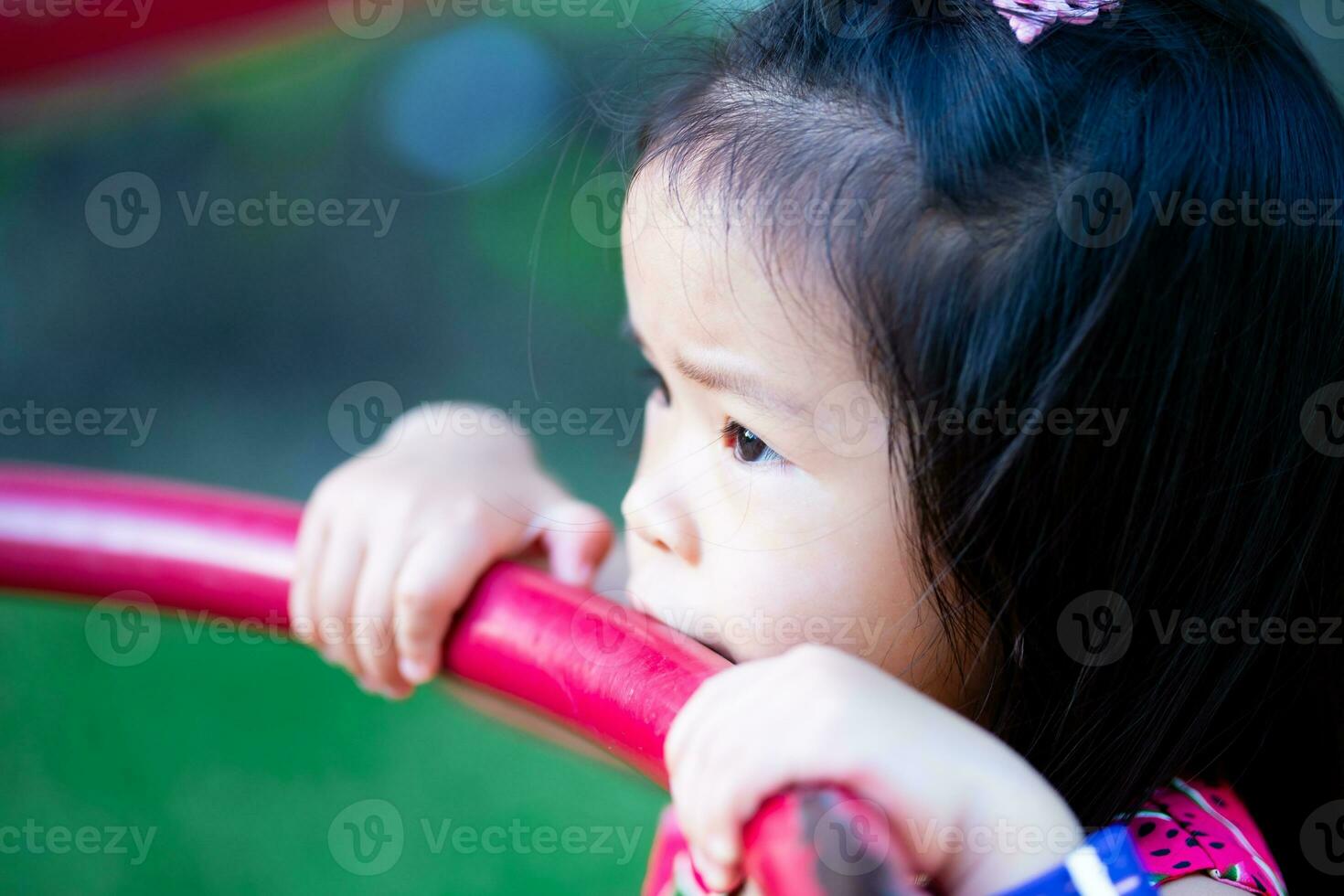 Closeup face of cute Asian girl using her mouth to touch object of playground equipment. Health care and cleanliness in young children. Empty space for entering text. photo