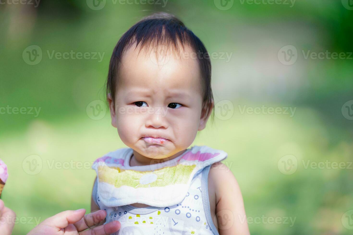 One year old Asian Baby Boy Makes a Serious Expression after Eating Sweet and Sour Strawberry Ice Cream, Child Wears an Apron, Toddler Eats Sweets. photo