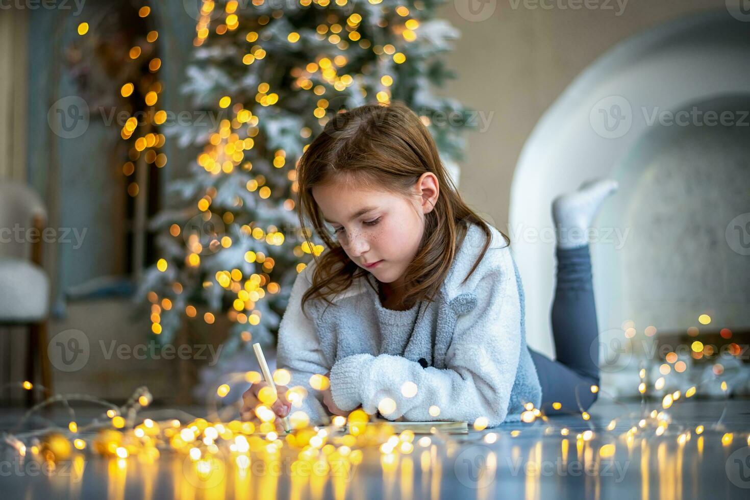 a cute girl lies on the floor near the Christmas tree and writes a letter to Santa Claus photo