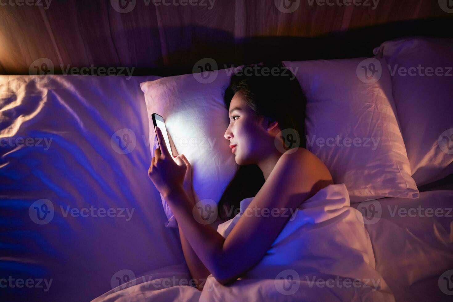 asiático mujer utilizando móvil teléfono teléfono inteligente tendido en el cama en el dormitorio. mujer desplazamiento social redes en móvil. contenido hembra acostado en cama debajo cobija y acecho película en teléfono inteligente foto