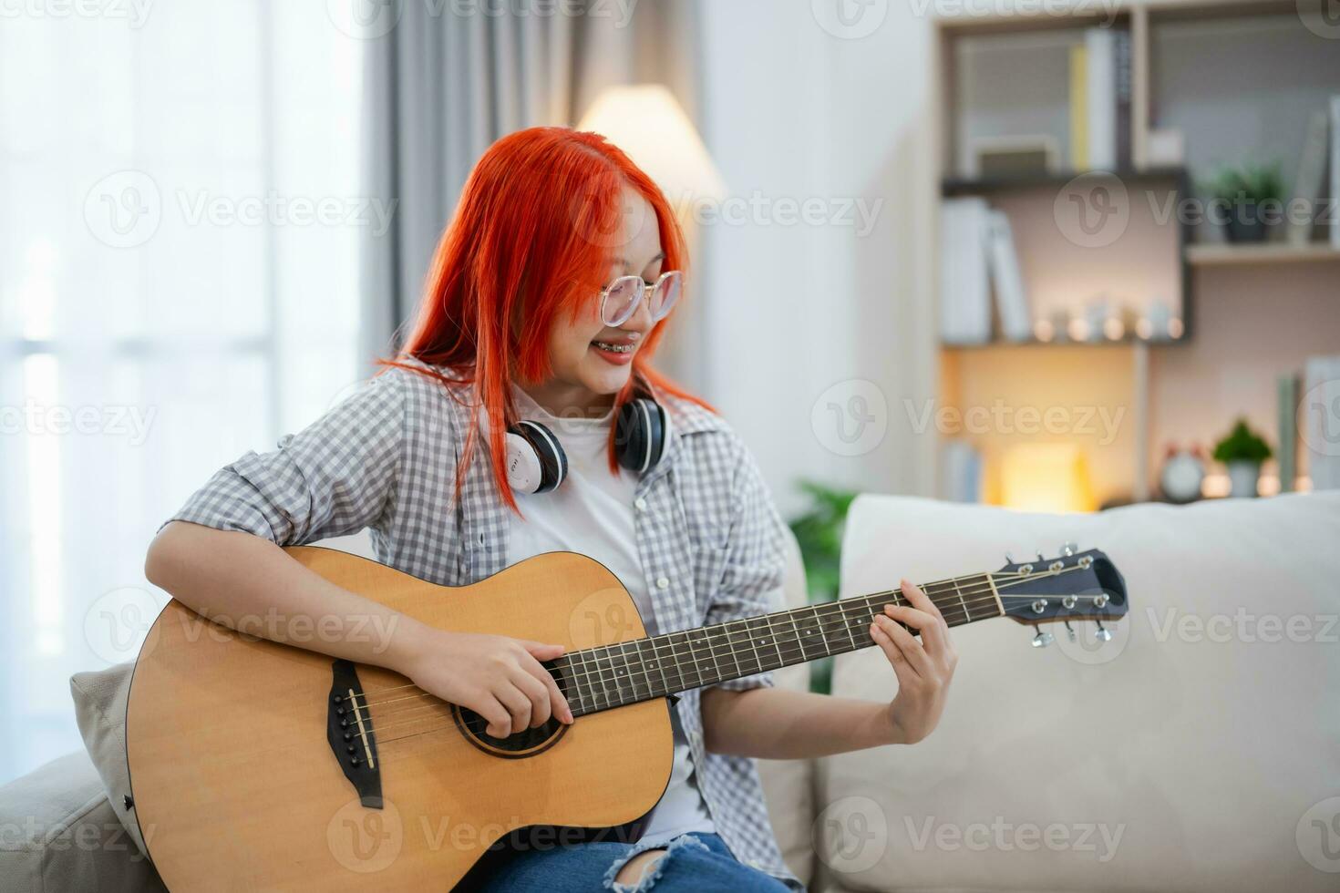 Asian woman wearing glasses and headphone playing guitar while sitting on sofa in the living room at home. Asian women writing song while playing guitar at home. Compose song music concept. photo