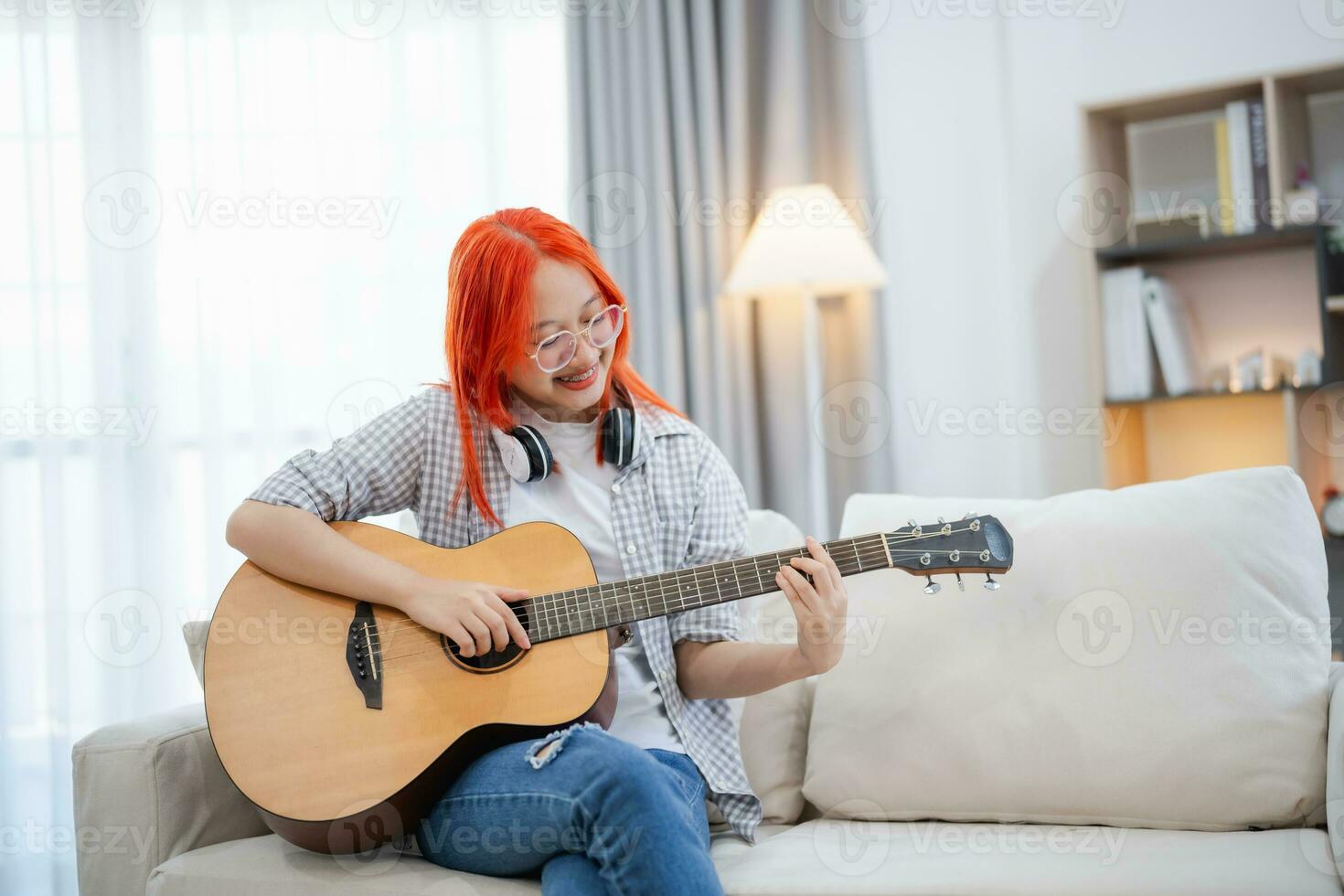 asiático mujer vistiendo lentes y auricular jugando guitarra mientras sentado en sofá en el vivo habitación a hogar. asiático mujer escritura canción mientras jugando guitarra a hogar. componer canción música concepto. foto