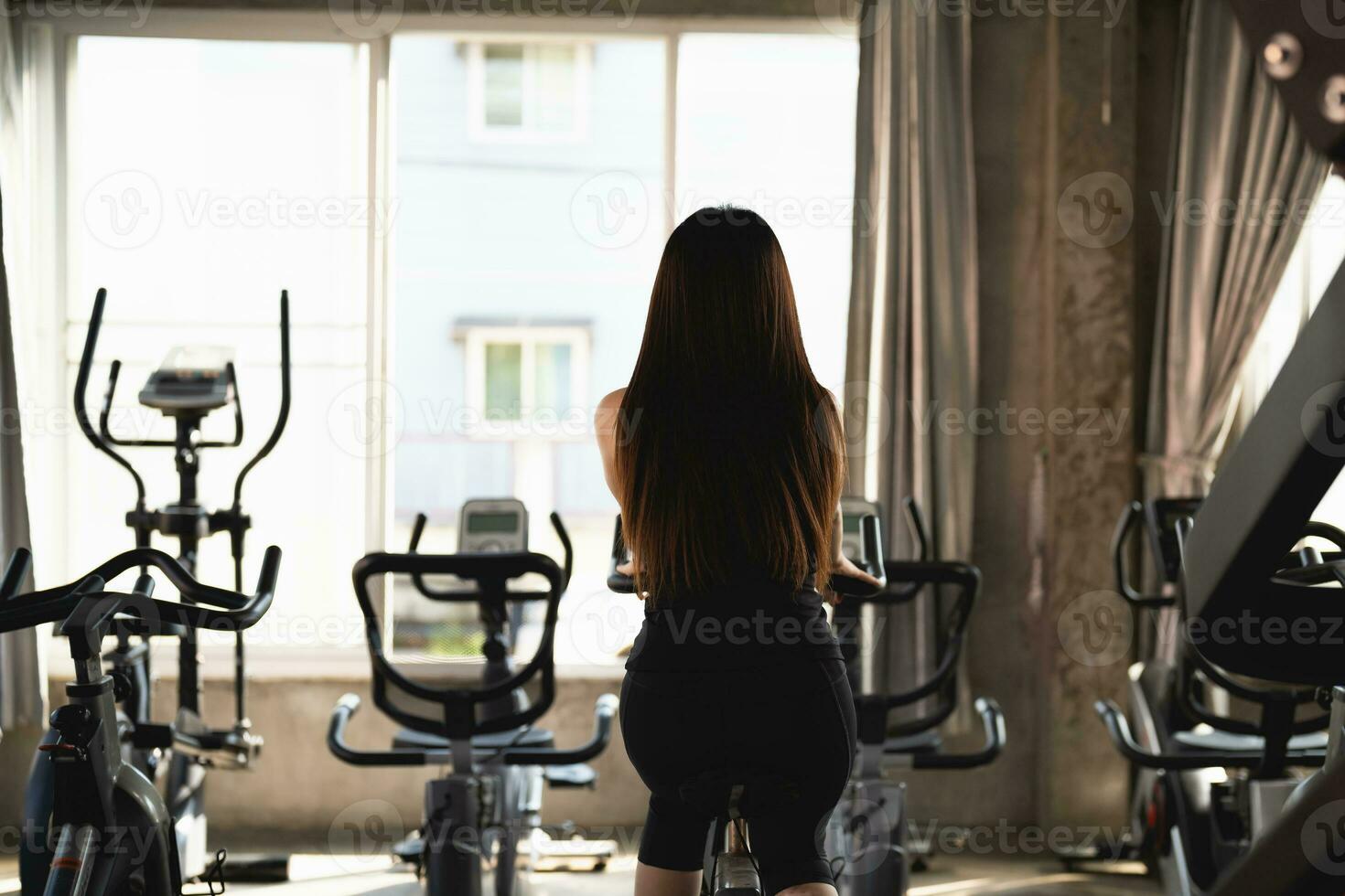 Back side of Asian sportswoman exercising on a bicycle in the gym, determination to cardio lose weight, makes her healthy. exercise bike woman fitness sport concept. photo