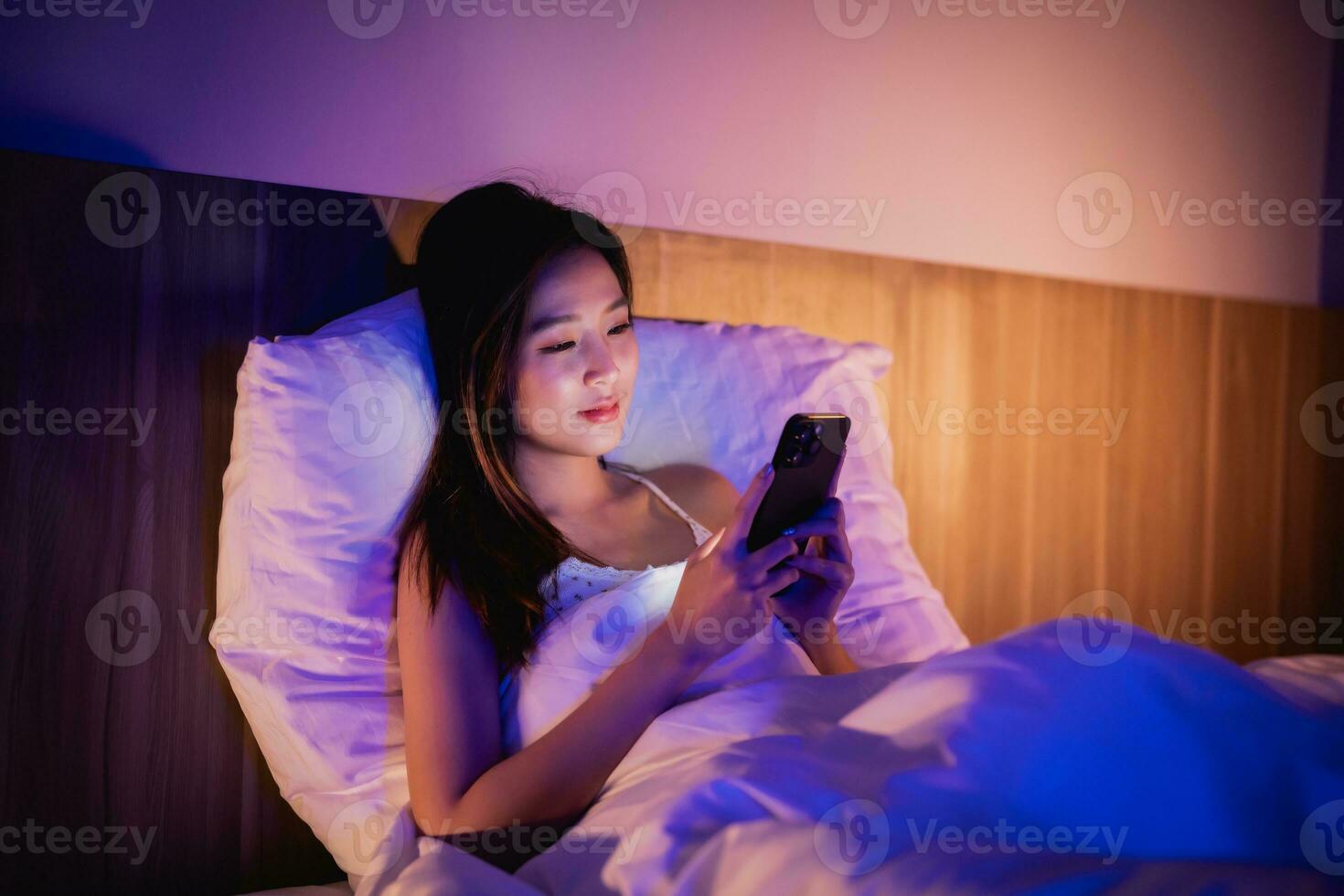 Asian woman using mobile phone smartphone laying on the bed in the bedroom. Women scrolling social networks on mobile. Content female lying in bed under blanket and watching movie on smartphone. photo