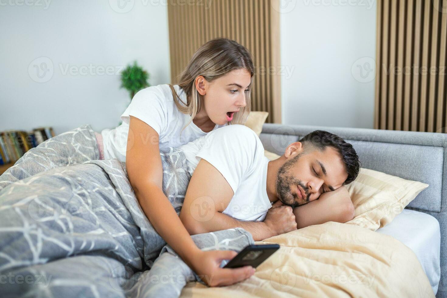 Woman is jealous and suspicious and spies in her partner's smartphone whiles he's sleeping in bedroom. The wife is spying on her husband's phone while he sleeps. The concept of distrust, jealousy photo