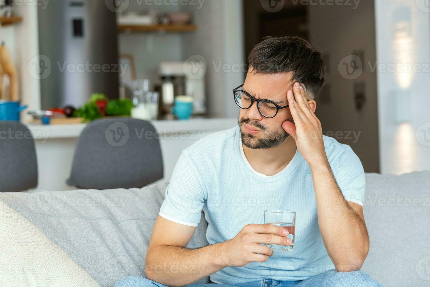 Young man suffering from strong headache or migraine sitting with glass of water on the sofa, millennial guy feeling intoxication and pain touching aching head, morning after hangover concept photo