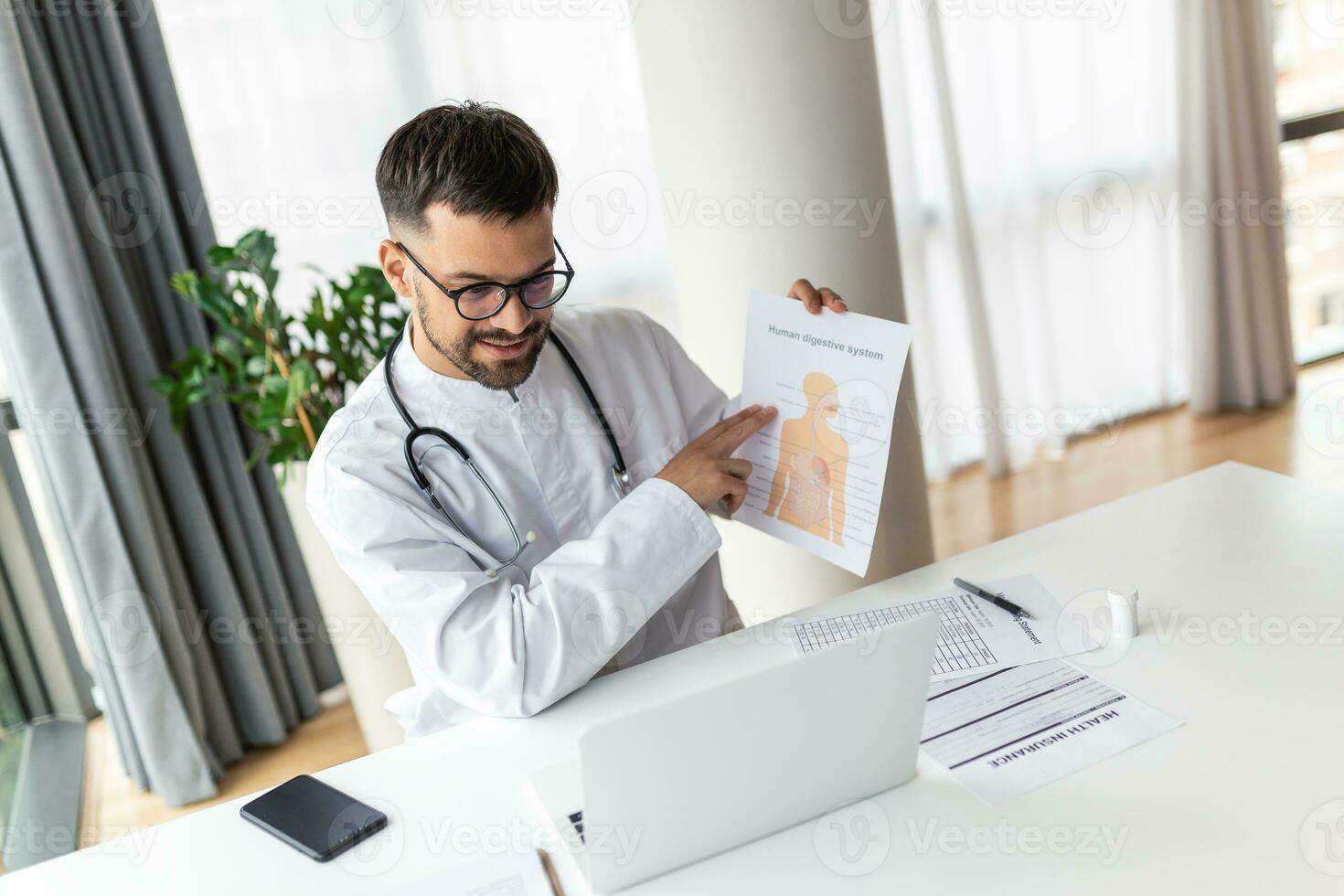 Doctor talking online with patient, showing him digestive system on clipboard, making video call, looking at camera, young male wearing white uniform with stethoscope speaking photo