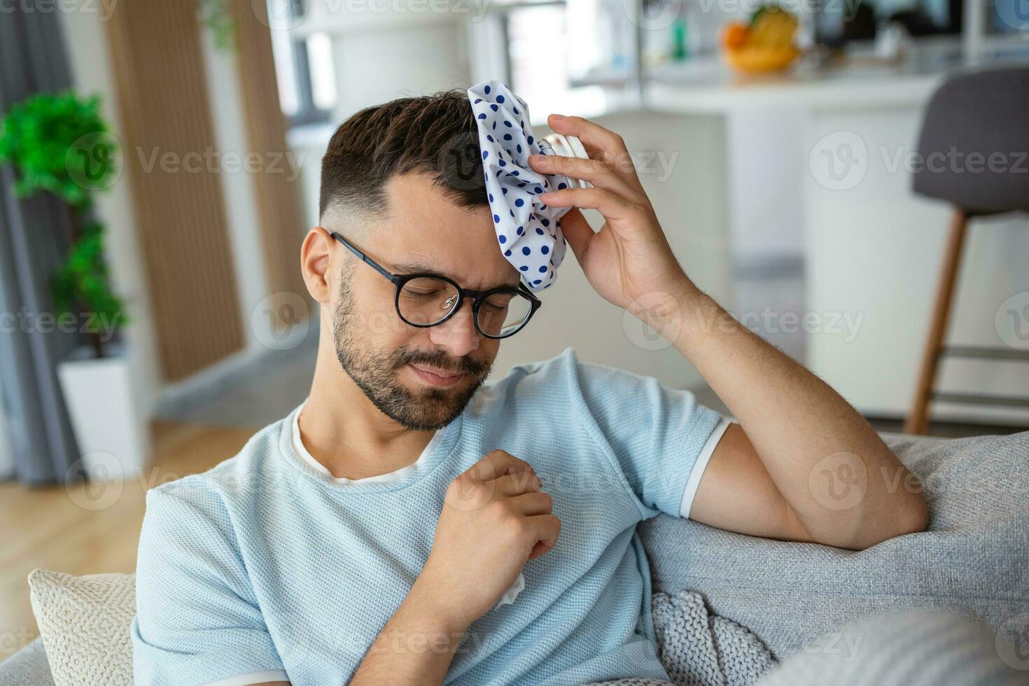Man feeling bad, sitting on the sofa with headache and applying ice bag photo