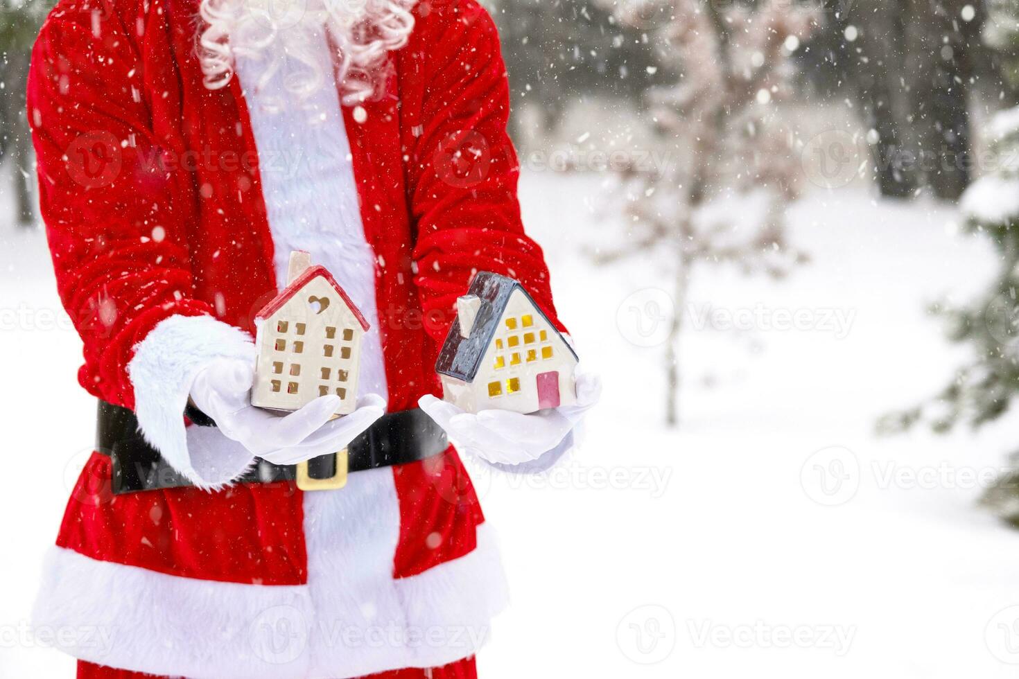 Papa Noel cuidadosamente sostiene en su manos un minúsculo casa en un pequeño Talla en nieve al aire libre - un regalo para Navidad, nuevo año, el cumplimiento de un sueño. proteccion, seguro, hipoteca, compra de alojamiento foto