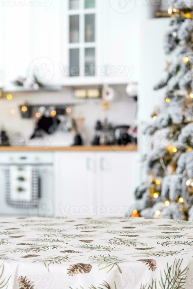 Round empty table to demonstrate the product with with tablecloth with pine needles and cones print, space for text in white kitchen, modern interior with a Christmas tree. New Year, Christmas photo