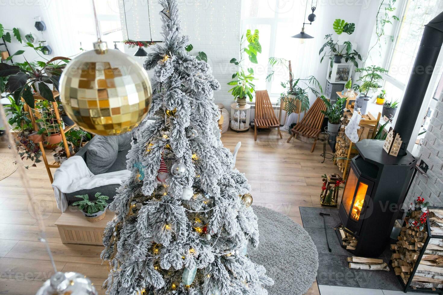 Festive white modern interior of house is decorated for Christmas and New Year in loft style with black stove, fireplace, Christmas tree. Potted plants, firewood in the woodpile photo