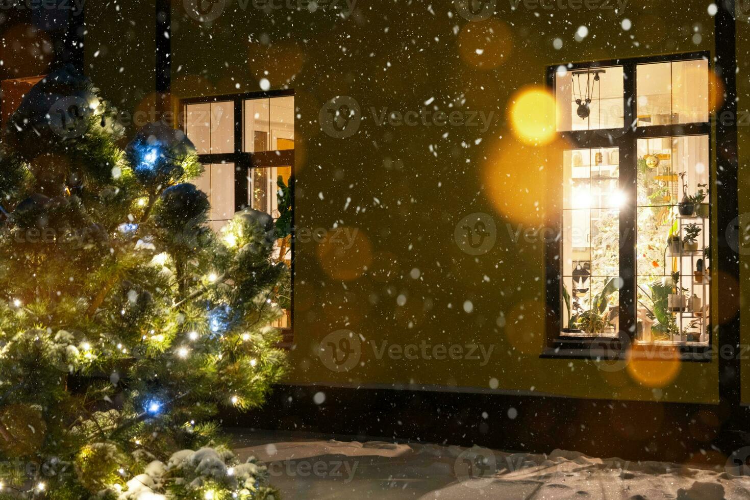 Cozy festive window of the house outside with the warm light of fairy lights garlands inside - celebrate Christmas and New Year in a warm home. Christmas tree, bokeh, snow on pine trees and snowfall photo