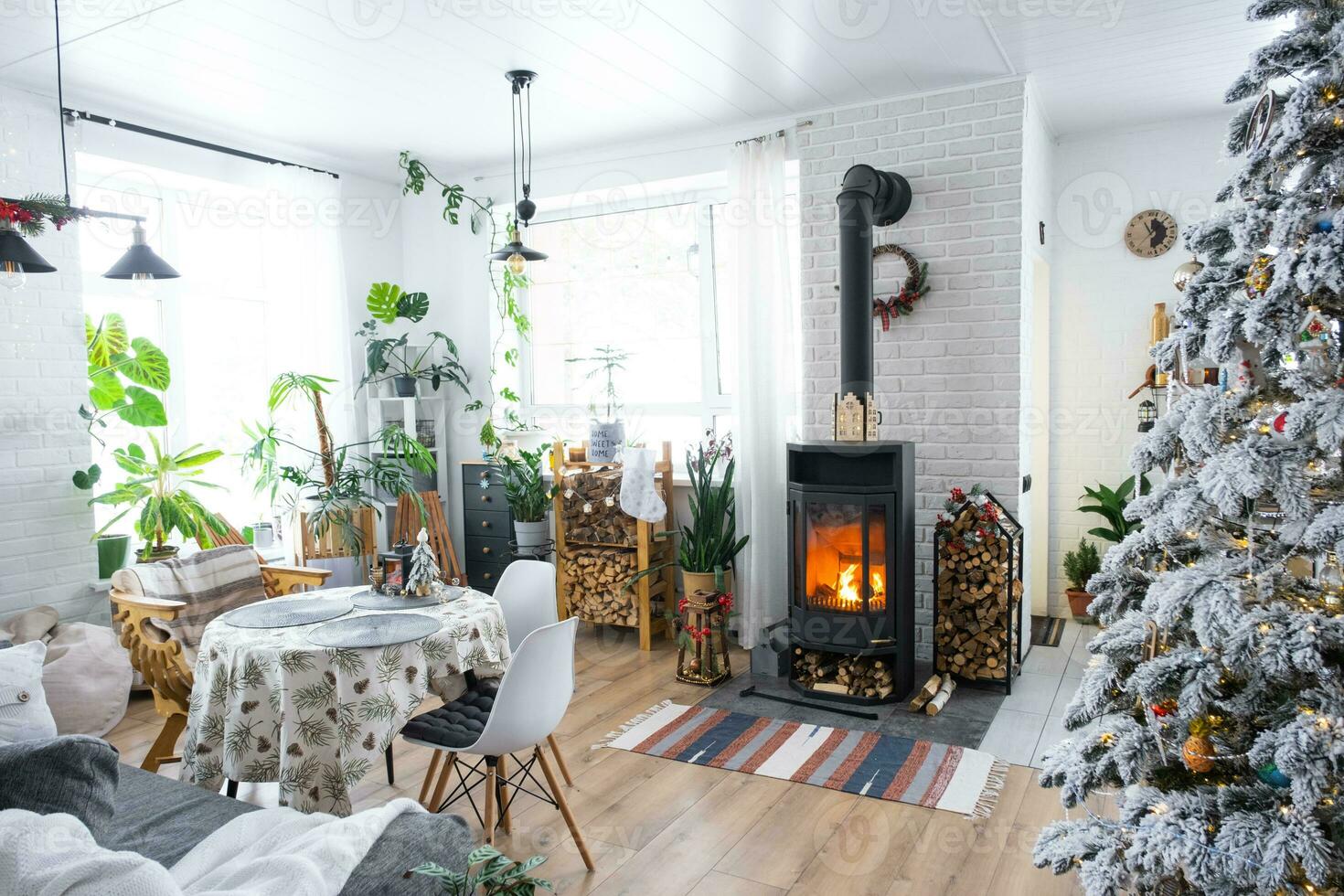 Festive white modern interior of house is decorated for Christmas and New Year in loft style with black stove, fireplace, Christmas tree. Potted plants, firewood in the woodpile photo
