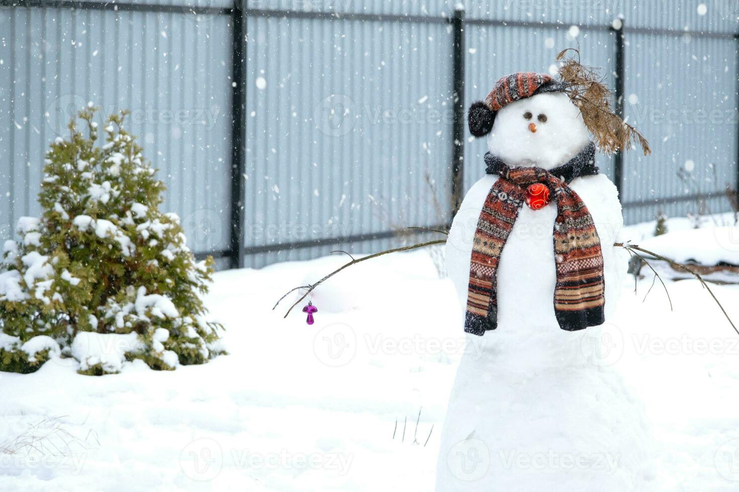 Funny snowman in a hat and scarf on the background of grey fence in the yard. Winter, winter entertainment, snowfall photo