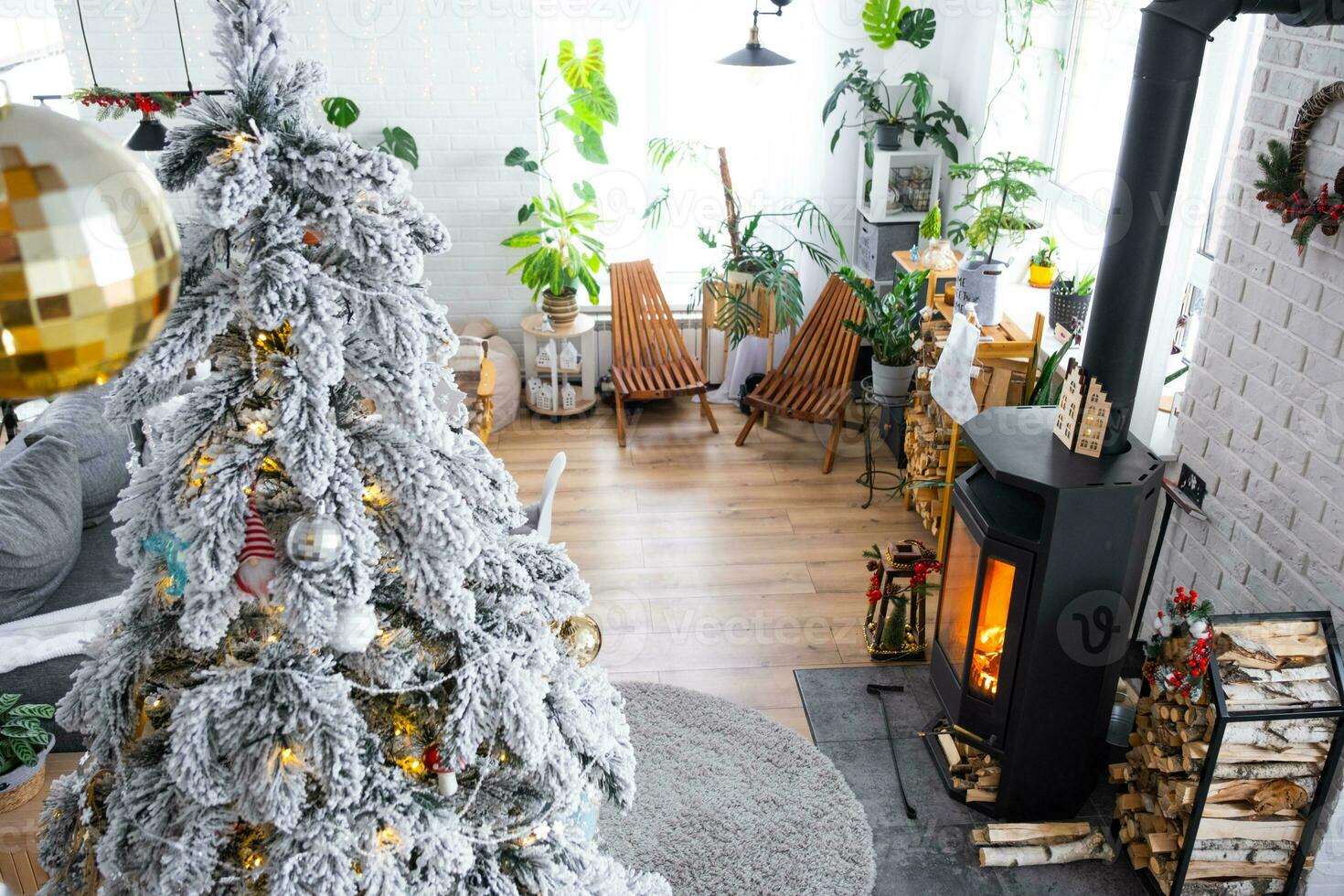 Festive white modern interior of house is decorated for Christmas and New Year in loft style with black stove, fireplace, Christmas tree. Potted plants, firewood in the woodpile photo