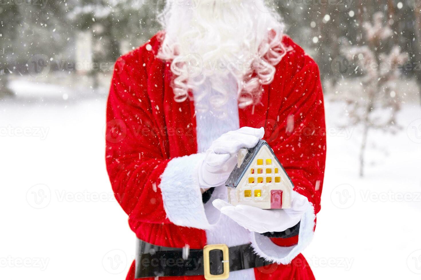 Papa Noel cuidadosamente sostiene en su manos un minúsculo casa en un pequeño Talla en nieve al aire libre - un regalo para Navidad, nuevo año, el cumplimiento de un sueño. proteccion, seguro, hipoteca, compra de alojamiento foto