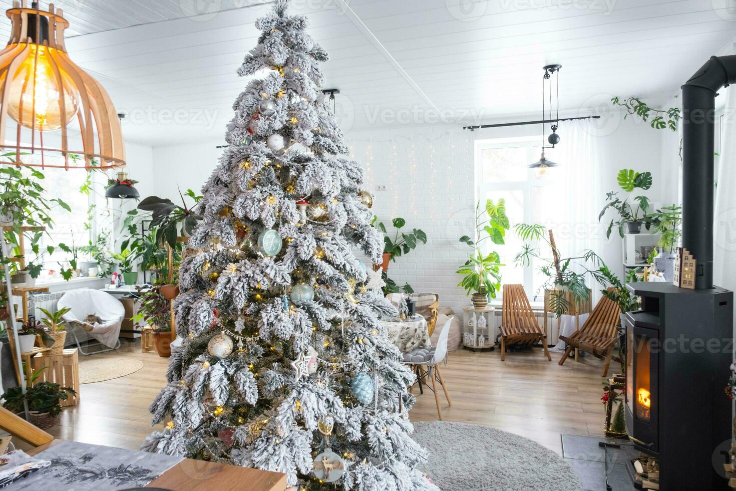 Festive white modern interior of house is decorated for Christmas and New Year in loft style with black stove, fireplace, Christmas tree. Potted plants, firewood in the woodpile photo