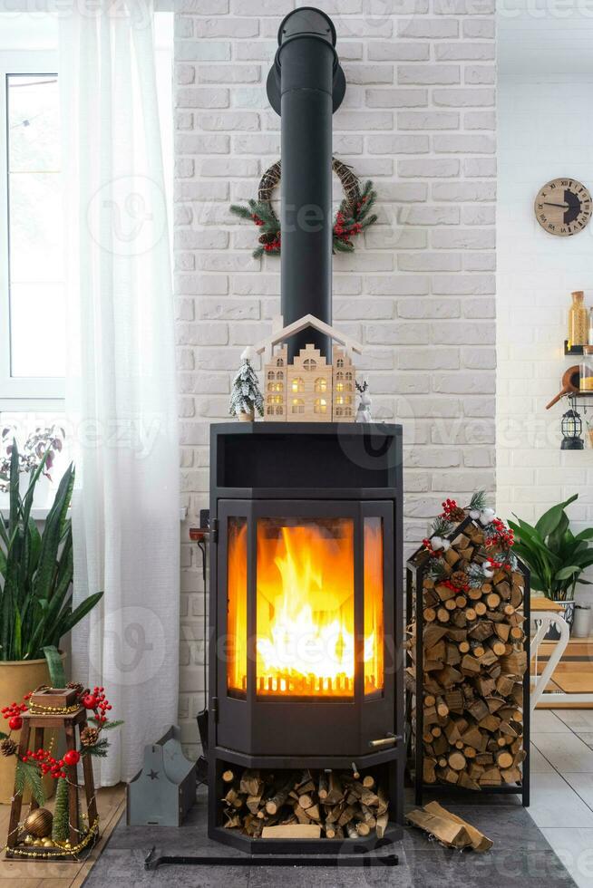 Metall black stove, burning hearth fireplace in white Festive interior of house is decorated for Christmas and New Year, Christmas tree. firewood in the woodpile, cozy and heating of home photo