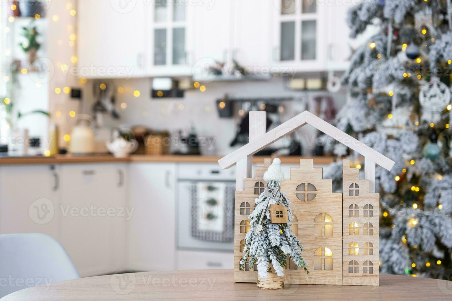 Key on Christmas tree and tiny house on cozy home with Christmas decor on table of festive white kitchen. Gift for New Year. Insurance, moving to new house, mortgage, rent and purchase real estate photo