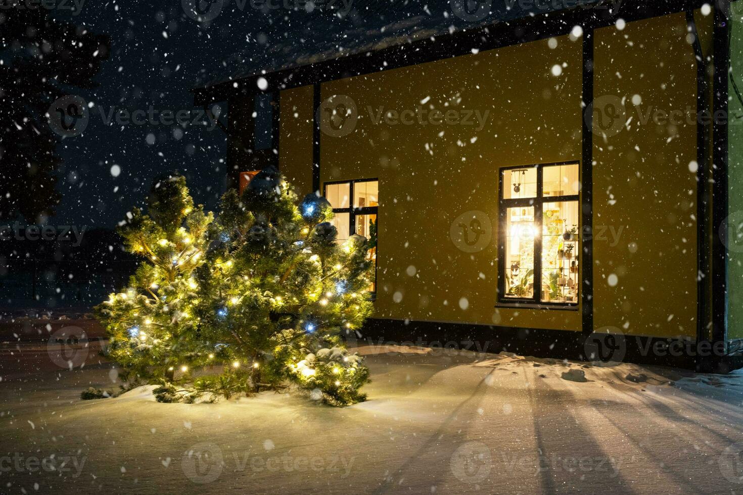 Cozy festive window of the house outside with the warm light of fairy lights garlands inside - celebrate Christmas and New Year in a warm home. Christmas tree, bokeh, snow on pine trees and snowfall photo
