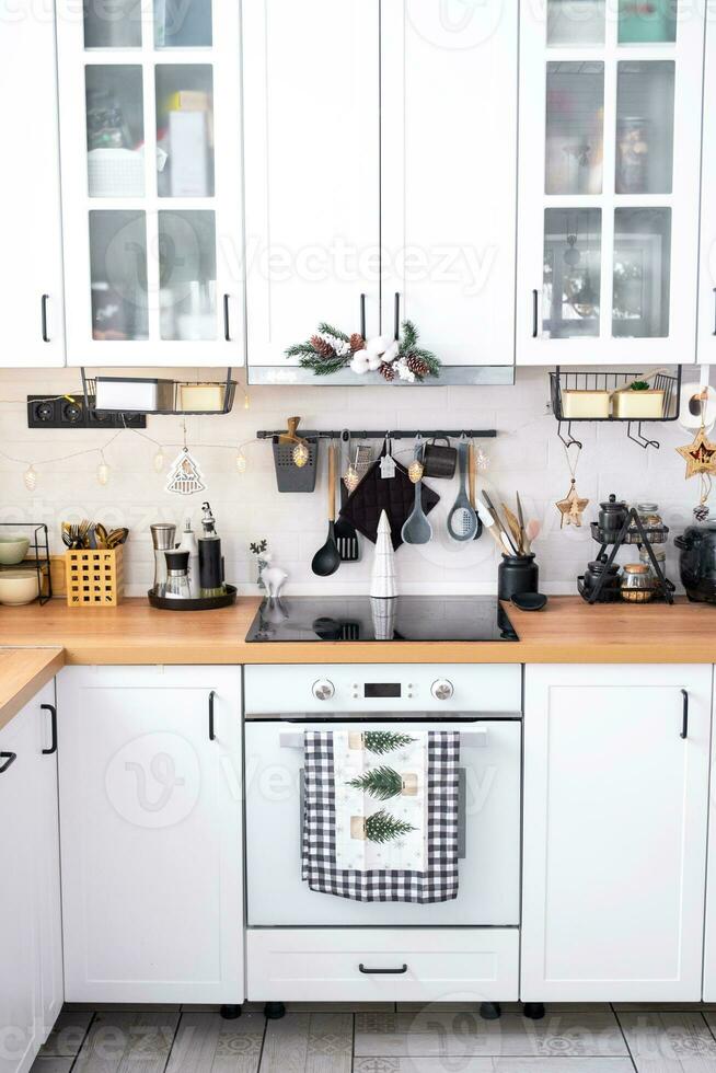 White Christmas decor in white kitchen, towel with Christmas trees, a branch of a fir tree on cabinets, induction cooker with oven. New Year, Christmas mood, cozy home. photo