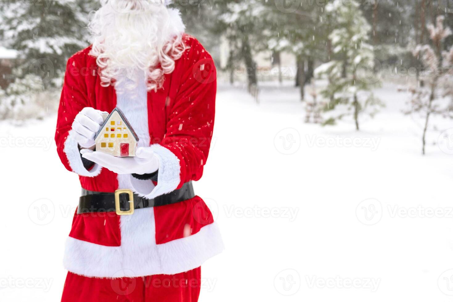 Santa carefully holds in his hands a tiny house in a small size in snow outdoor - a gift for Christmas, New year, the fulfillment of a dream. Protection, insurance, mortgage, purchase of housing photo