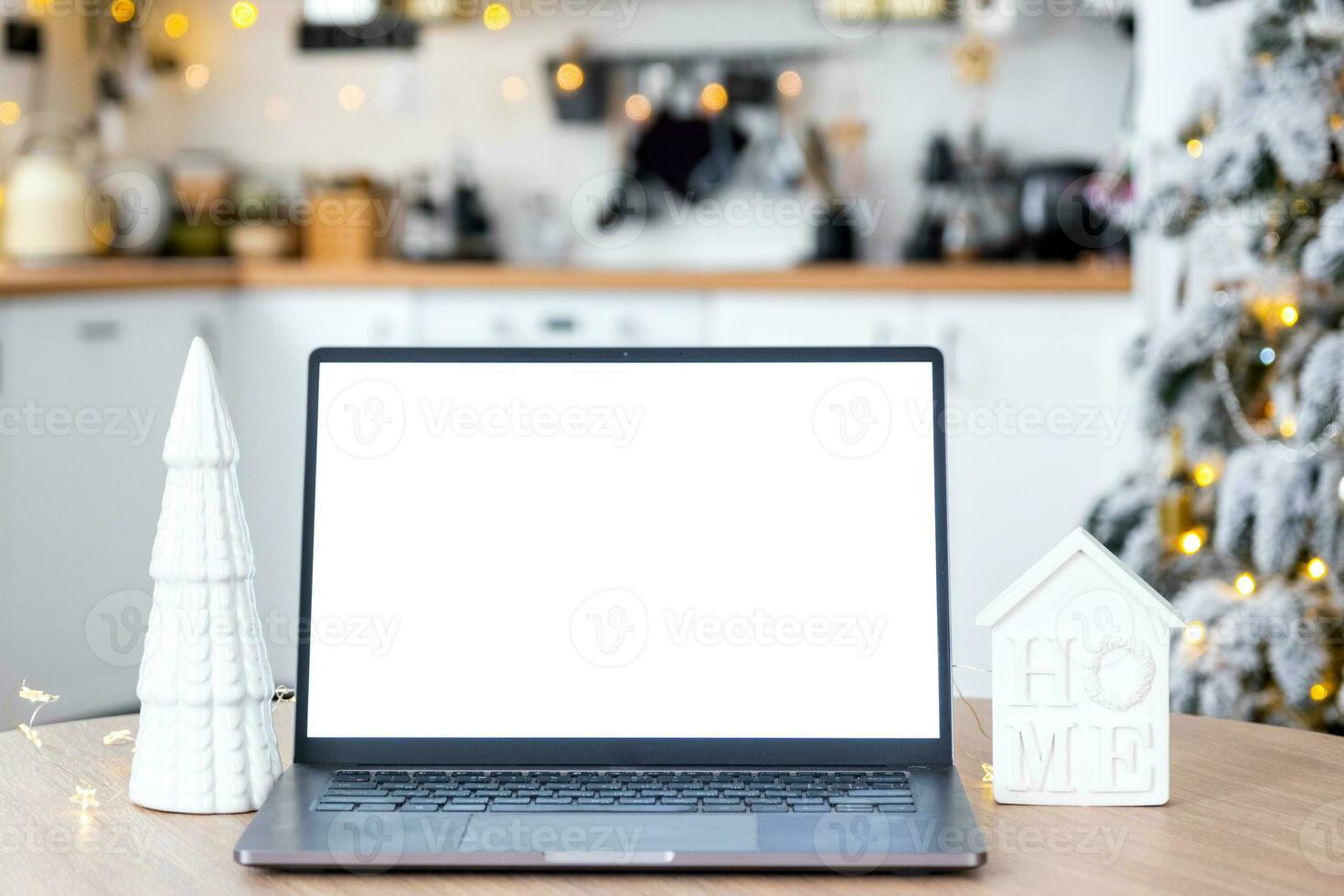 Laptop with a white screen mock up in the cozy white decorated Christmas kitchen with fairy lights and a Christmas tree. Seasonal remote work, internet, shopping, buying gifts Christmas and New Year. photo