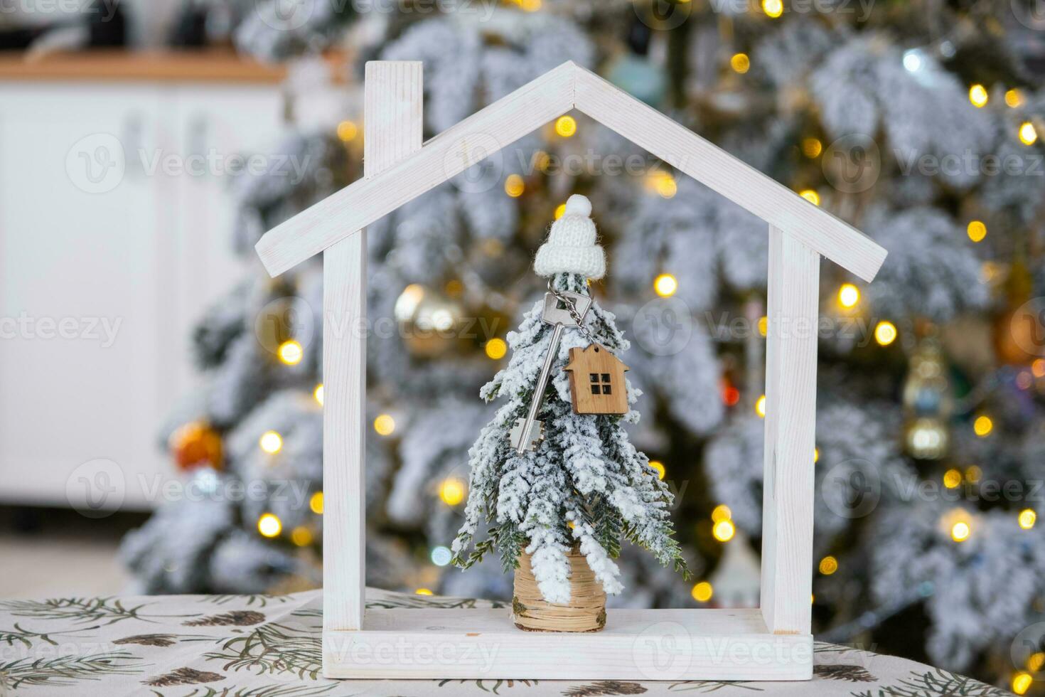 Key on Christmas tree and tiny house on cozy home with Christmas decor on table of festive white kitchen. Gift for New Year. Insurance, moving to new house, mortgage, rent and real estate photo