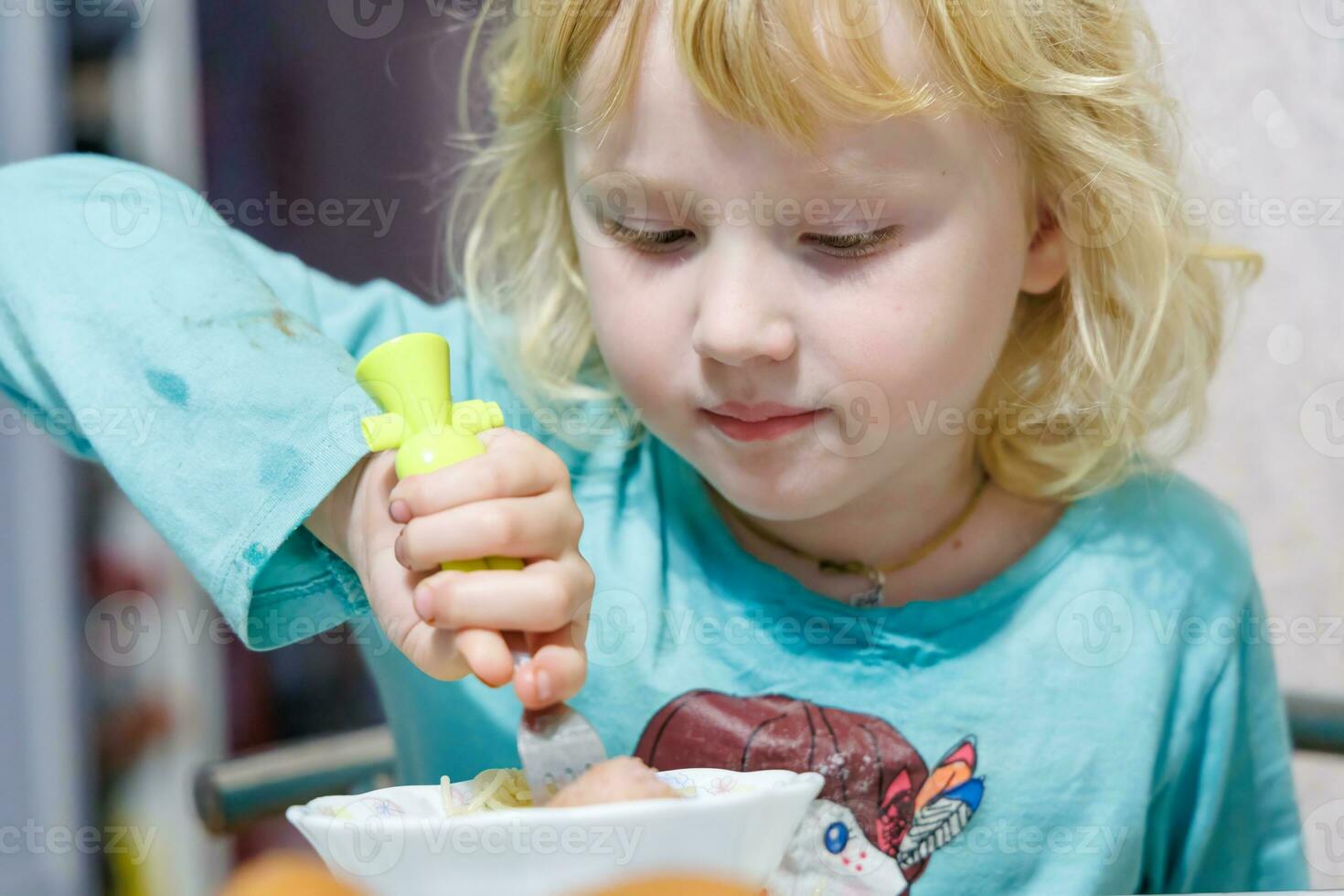 A little girl has breakfast at home spaghetti with sausages. Little blonde girl eating dinner with fork at table photo