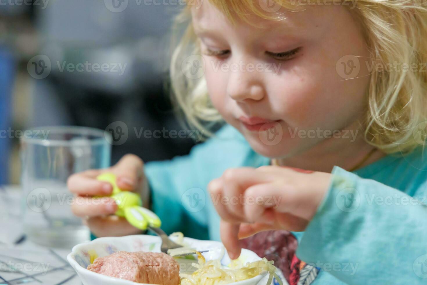 A little girl has breakfast at home spaghetti with sausages. Little blonde girl eating dinner with fork at table photo