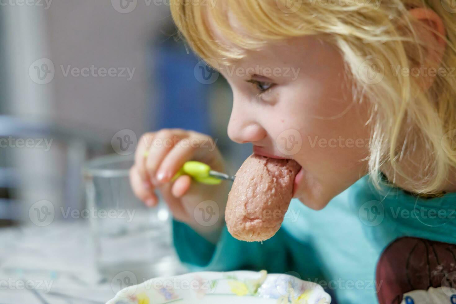 un pequeño niña tiene desayuno a hogar espaguetis con salchichas pequeño rubia niña comiendo cena con tenedor a mesa foto