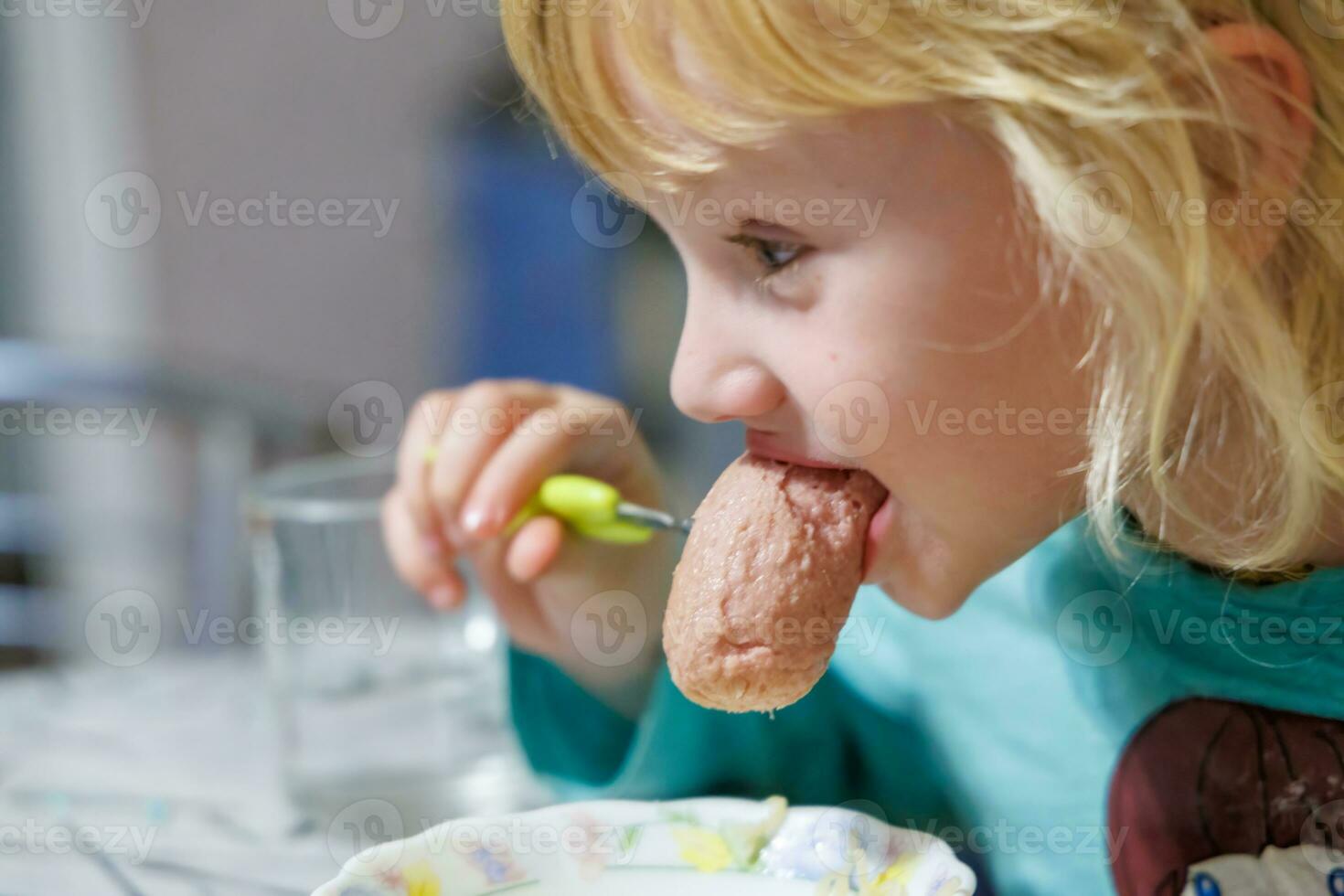 A little girl has breakfast at home spaghetti with sausages. Little blonde girl eating dinner with fork at table photo