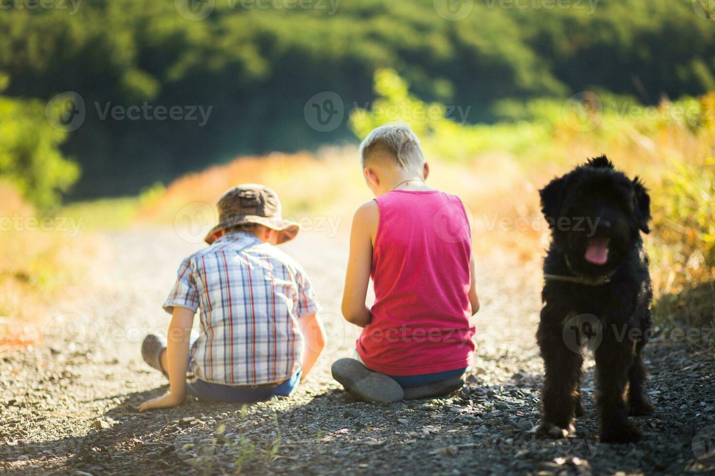 Two boys and a black dog sitting on the road in the forest photo