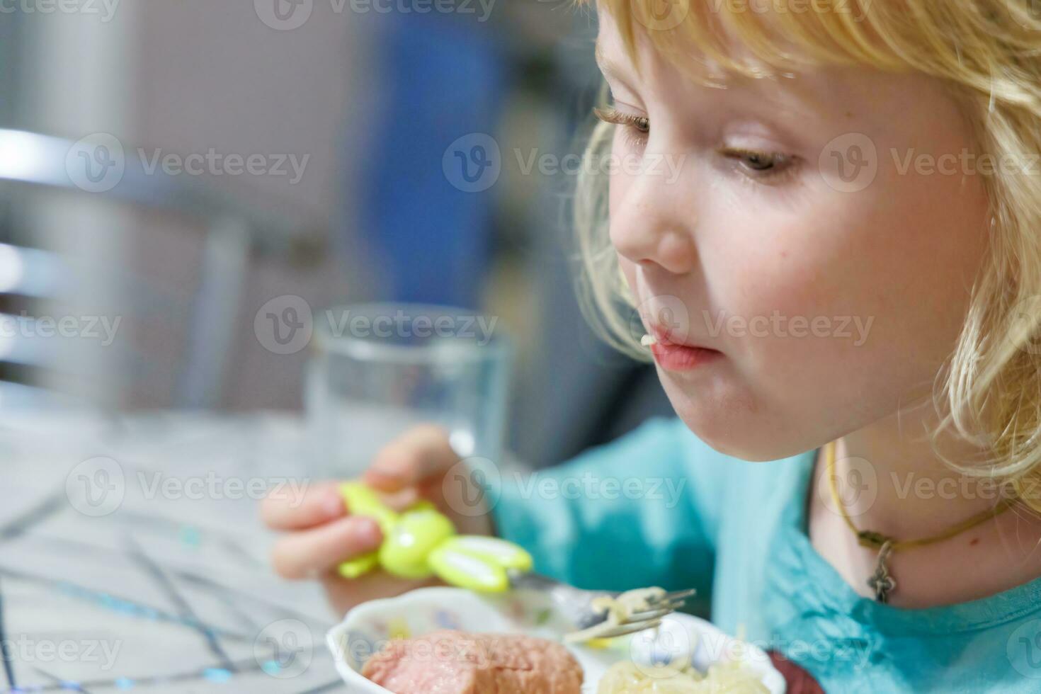 un pequeño niña tiene desayuno a hogar espaguetis con salchichas pequeño rubia niña comiendo cena con tenedor a mesa foto