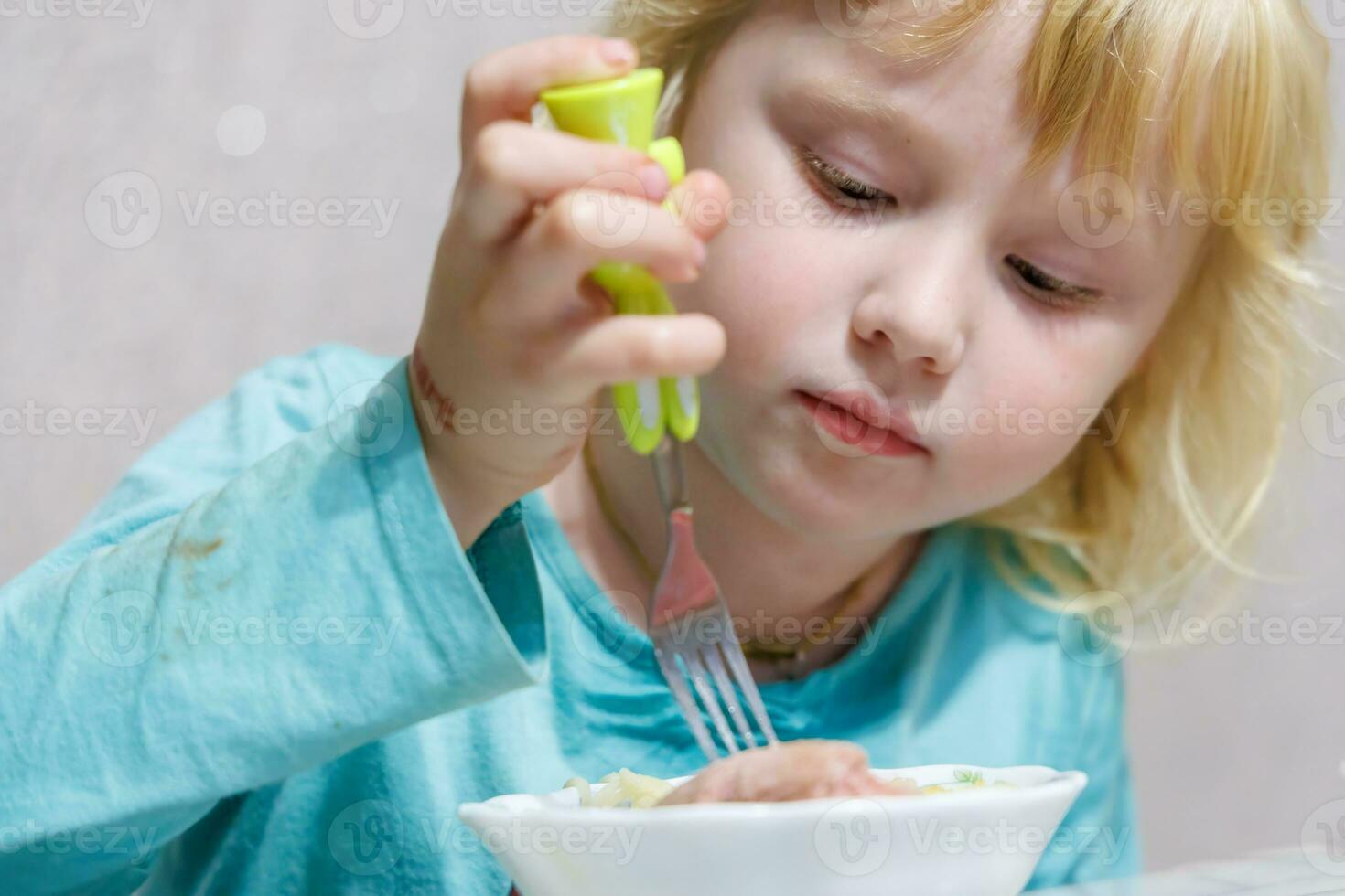 un pequeño niña tiene desayuno a hogar espaguetis con salchichas pequeño rubia niña comiendo cena con tenedor a mesa foto