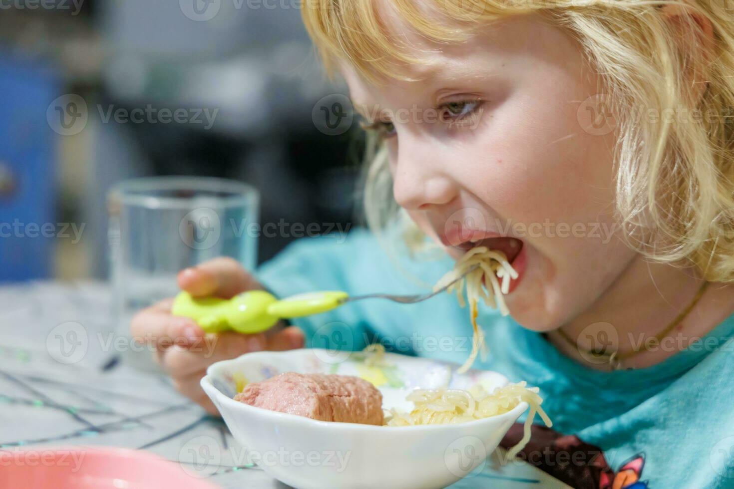 un pequeño niña tiene desayuno a hogar espaguetis con salchichas pequeño rubia niña comiendo cena con tenedor a mesa foto