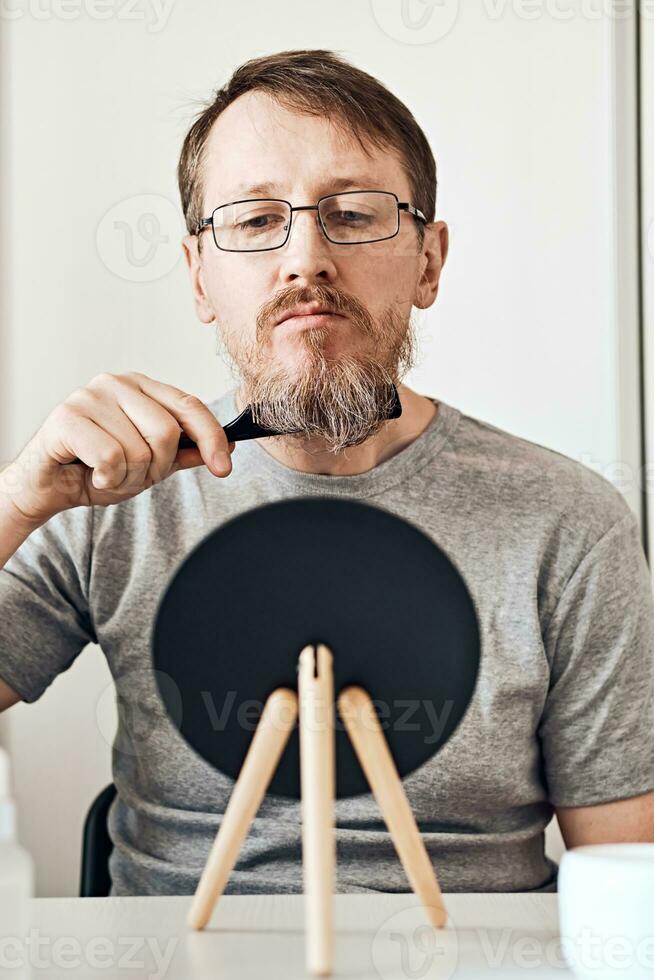 An attractive middle-aged man straightens his beard in front of a mirror. Natural look, light blond hair and freckles, beard with gray hair. Soft focus photo