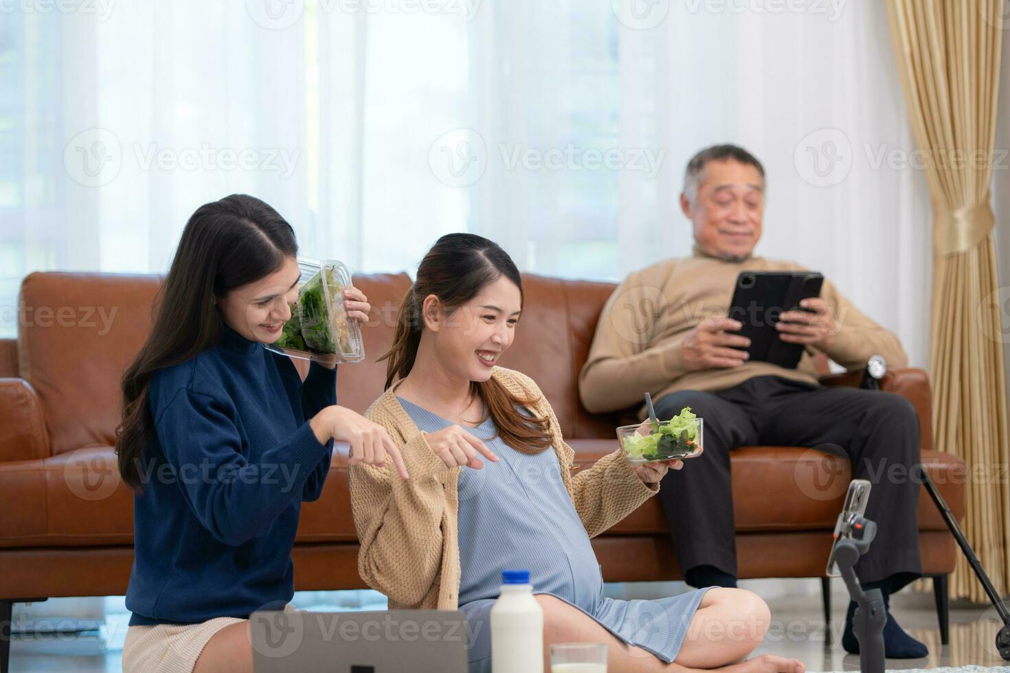 A pregnant asian woman sits on the floor with her younger sister. Live social media broadcast selling pregnant mothers' products and meals, There is a father in the background relaxing on a sofa. photo