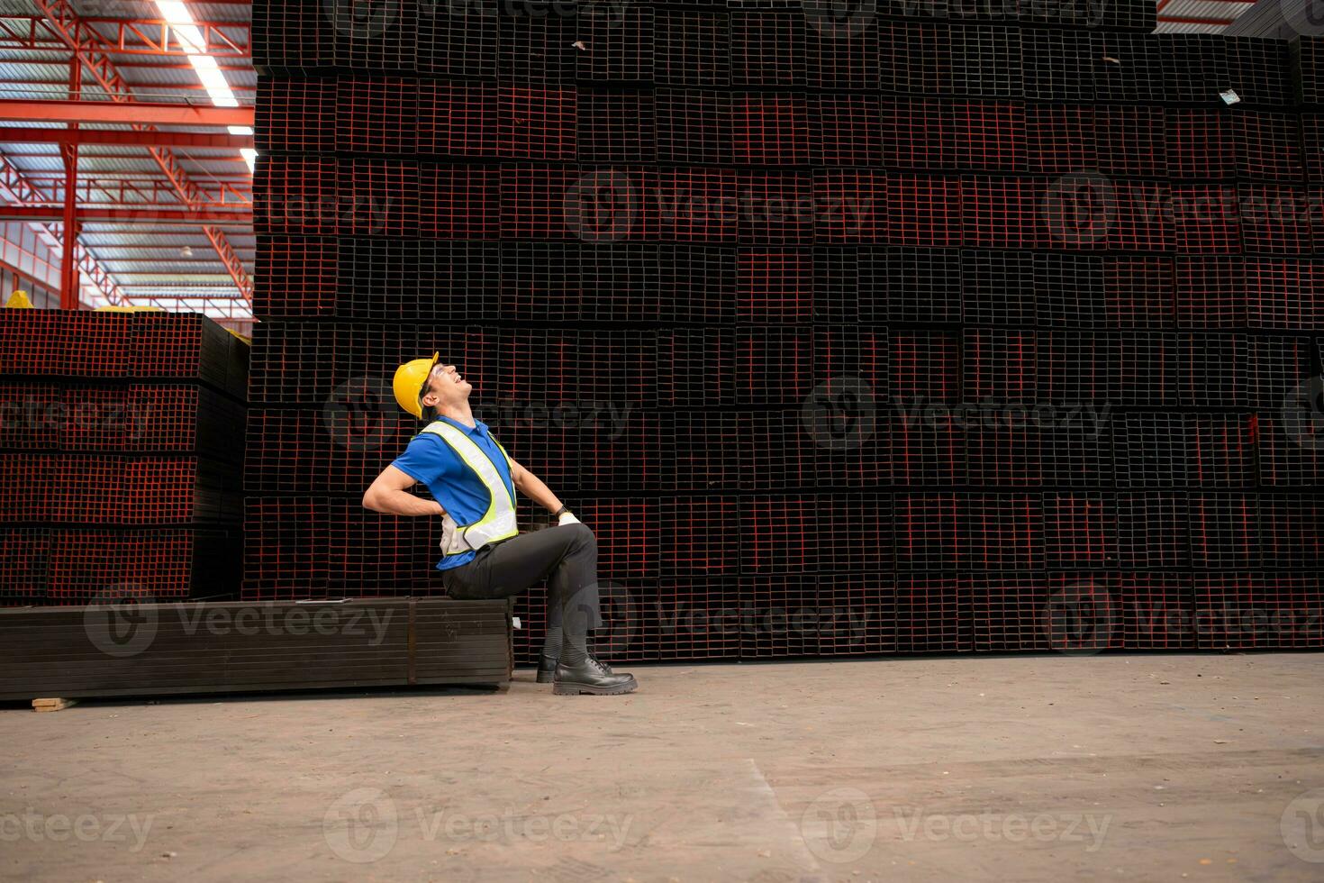 Portrait of a male worker wearing a safety vest and helmet sitting on a steels pallet due to back pain from working in a factory lifting heavy things. photo