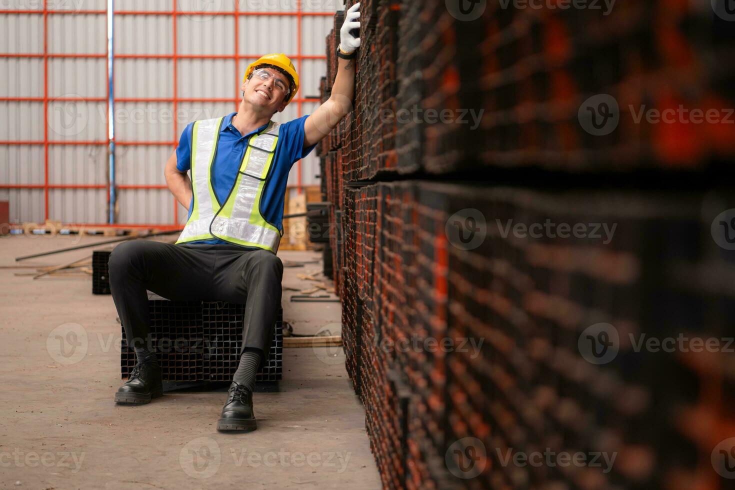 retrato de un masculino trabajador vistiendo un la seguridad chaleco y casco sentado en un aceros paleta debido a espalda dolor desde trabajando en un fábrica levantamiento pesado cosas. foto