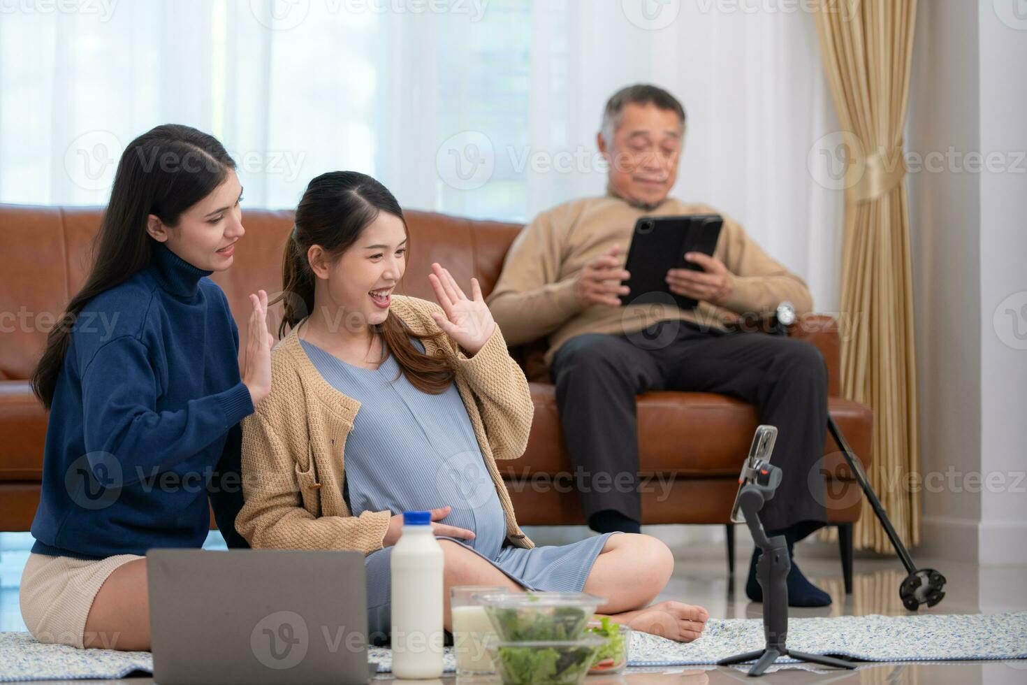 A pregnant asian woman sits on the floor with her younger sister. Live social media broadcast selling pregnant mothers' products and meals, There is a father in the background relaxing on a sofa. photo