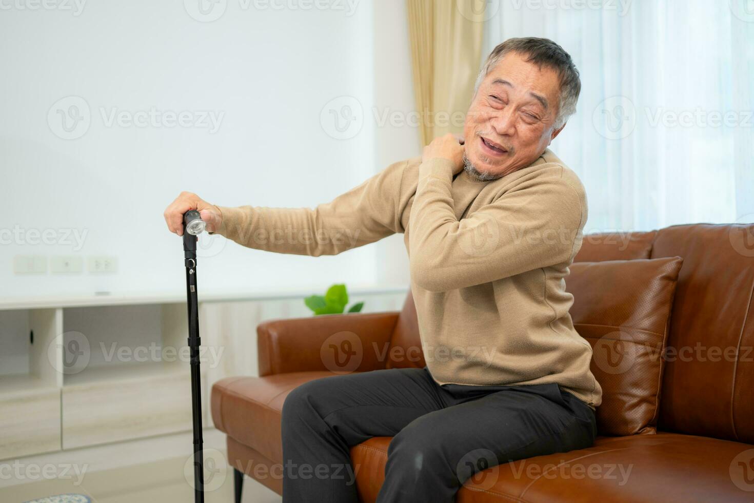 With shoulder pain, a senior guy sits on a sofa with a walking stick in his living room at home. photo