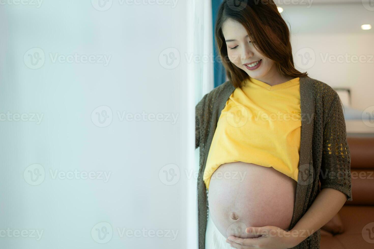 Beautiful pregnant woman standing near window at home and holding her belly photo