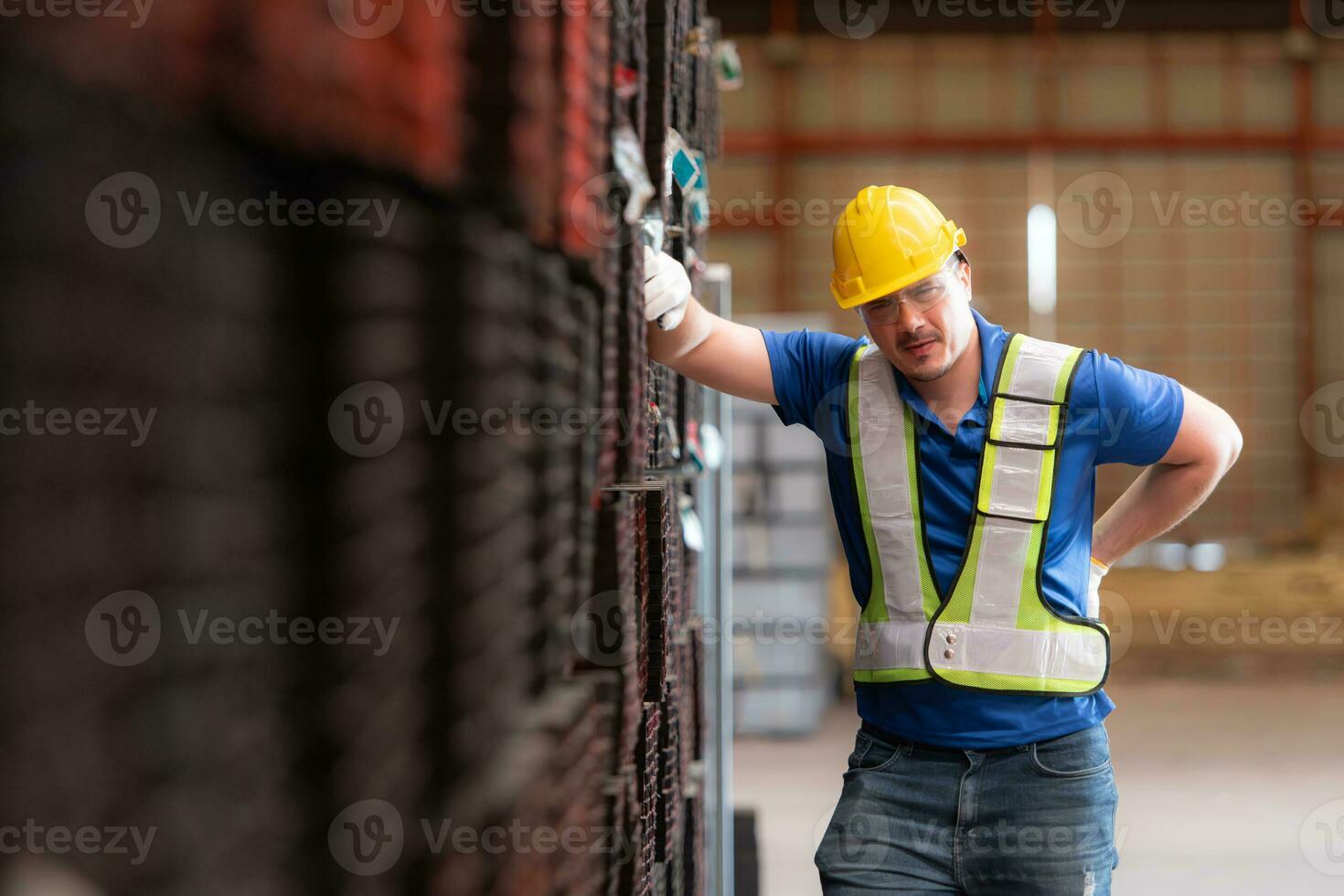 retrato de un masculino trabajador vistiendo un la seguridad chaleco y casco en pie en un aceros paleta debido a espalda dolor desde trabajando en un fábrica levantamiento pesado cosas. foto