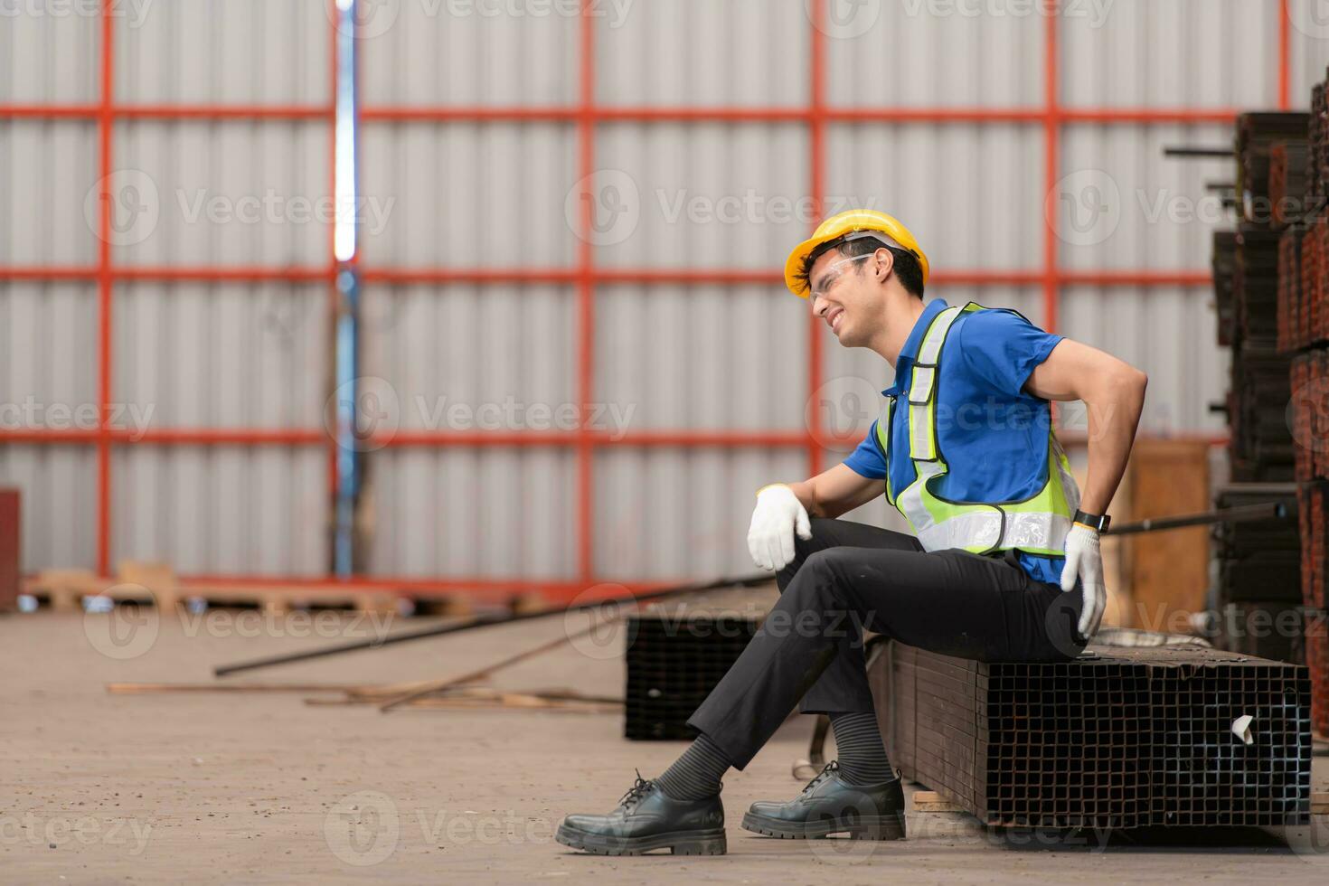 Portrait of a male worker wearing a safety vest and helmet sitting on a steels pallet due to back pain from working in a factory lifting heavy things. photo