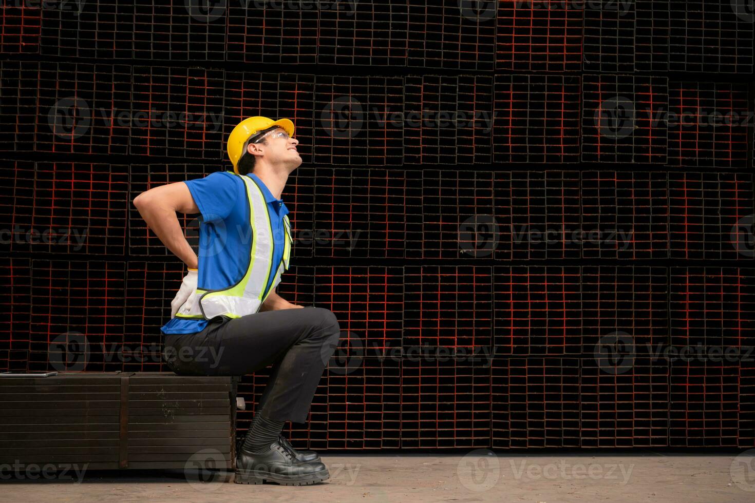 retrato de un masculino trabajador vistiendo un la seguridad chaleco y casco sentado en un aceros paleta debido a espalda dolor desde trabajando en un fábrica levantamiento pesado cosas. foto