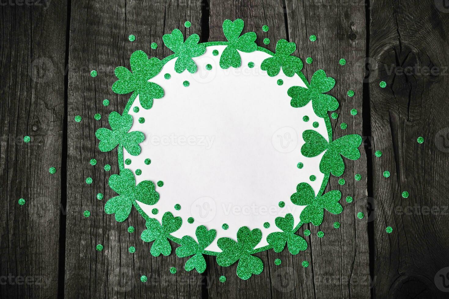 St. Patricks Day background. Round frame of shiny shamrocks and confetti on rough wooden background photo