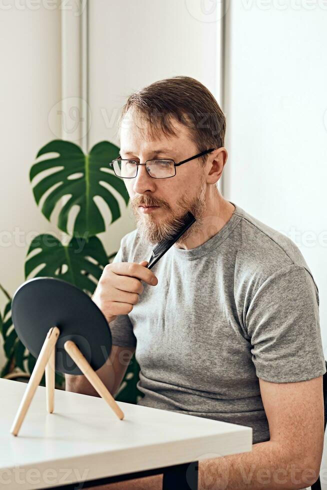 An attractive middle-aged man straightens his beard in front of a mirror. Natural look, light blond hair and freckles, beard with gray hair. Soft focus photo